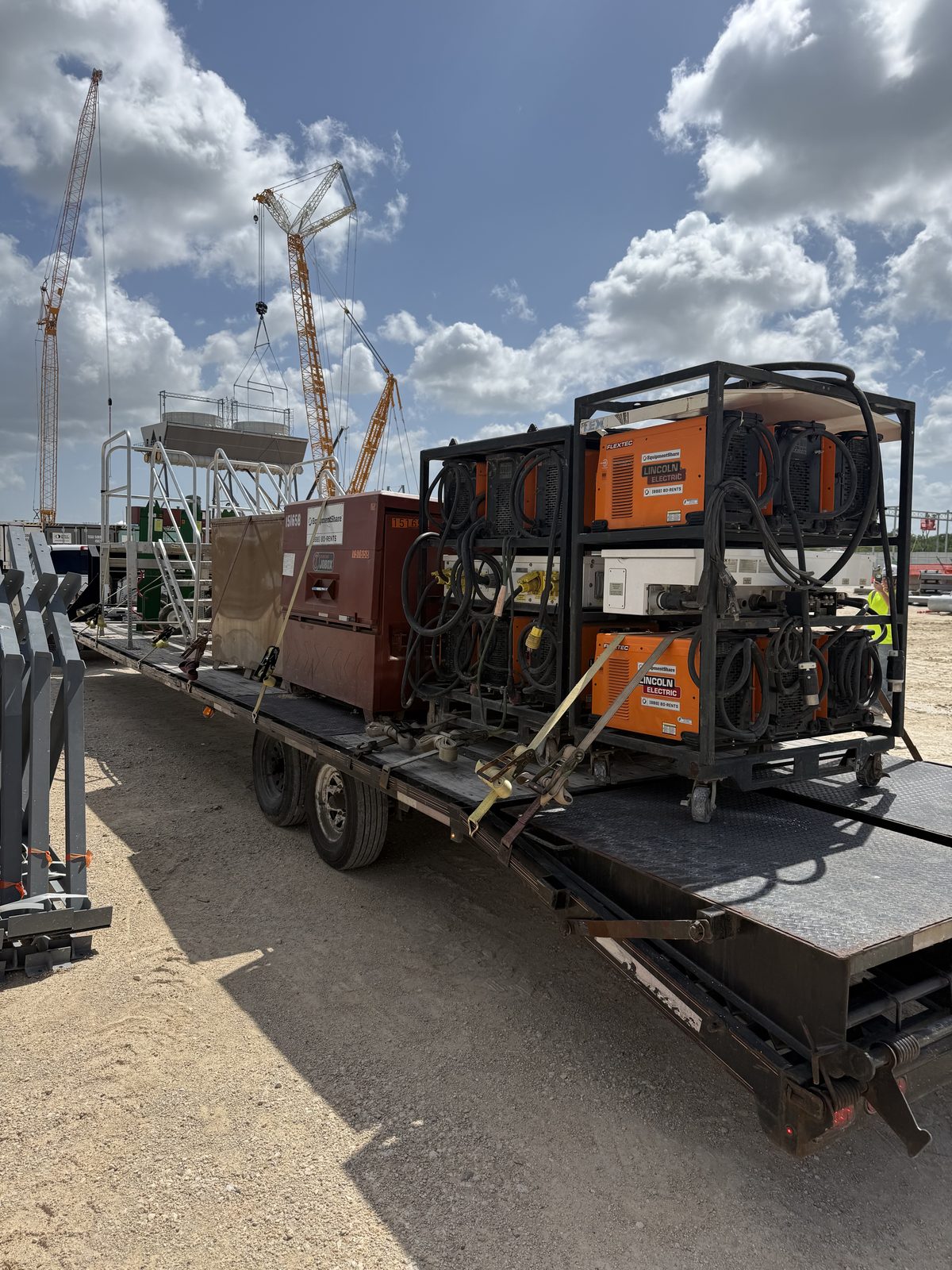 Step-deck trailer loaded with racked welding machines at a construction site, cranes in the background