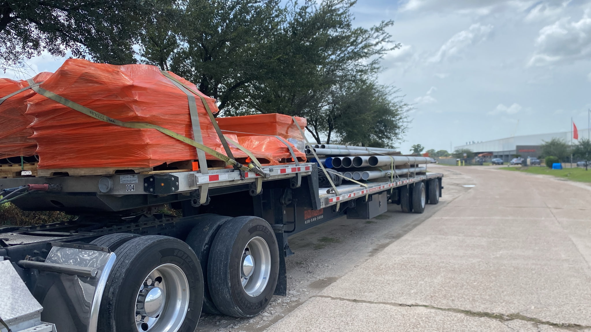 Flatbed trailer loaded with orange-wrapped palletized freight, a second flatbed of pipe behind it