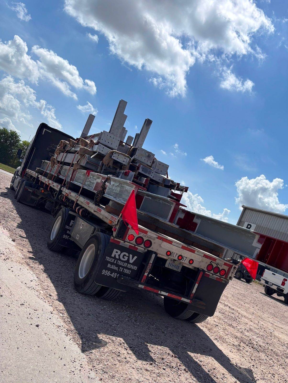 Flatbed trailer loaded with structural steel beams, red flag marking the overhang, with a red pipe trailer behind it
