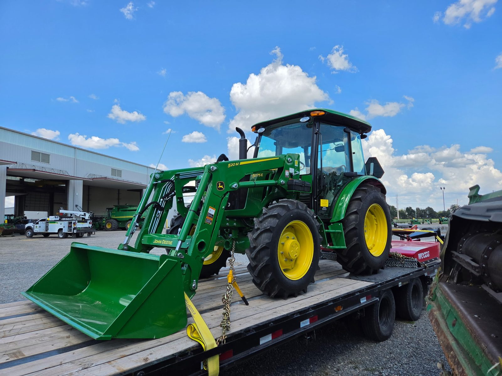 New John Deere tractor with a front loader chained down on a wood-deck flatbed trailer at an equipment dealership