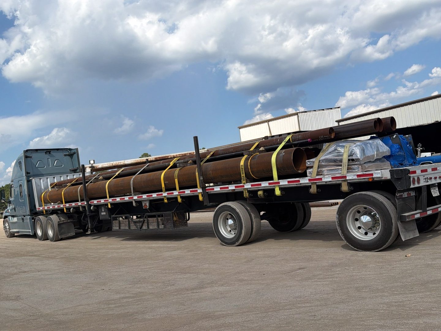Flatbed trailer loaded with large-diameter steel pipe and shrink-wrapped pallets, strapped down at a pipe yard