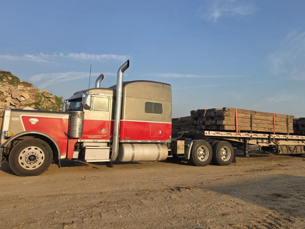 Peterbilt tractor and flatbed trailer loaded with stacked heavy timber, strapped down at a yard