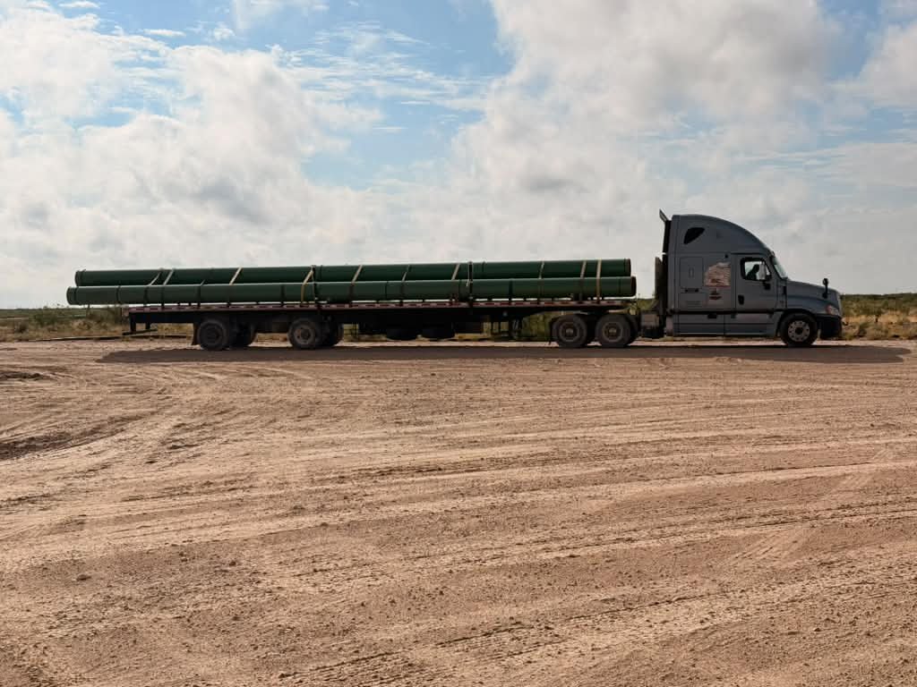 Flatbed trailer loaded with green coated large-diameter line pipe on a caliche pad in West Texas