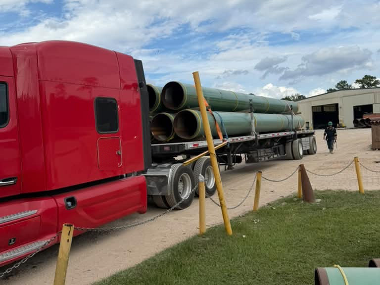 Red sleeper tractor coupled to a flatbed trailer loaded with green coated large-diameter line pipe at a pipe yard, stacked pipe and a warehouse behind it