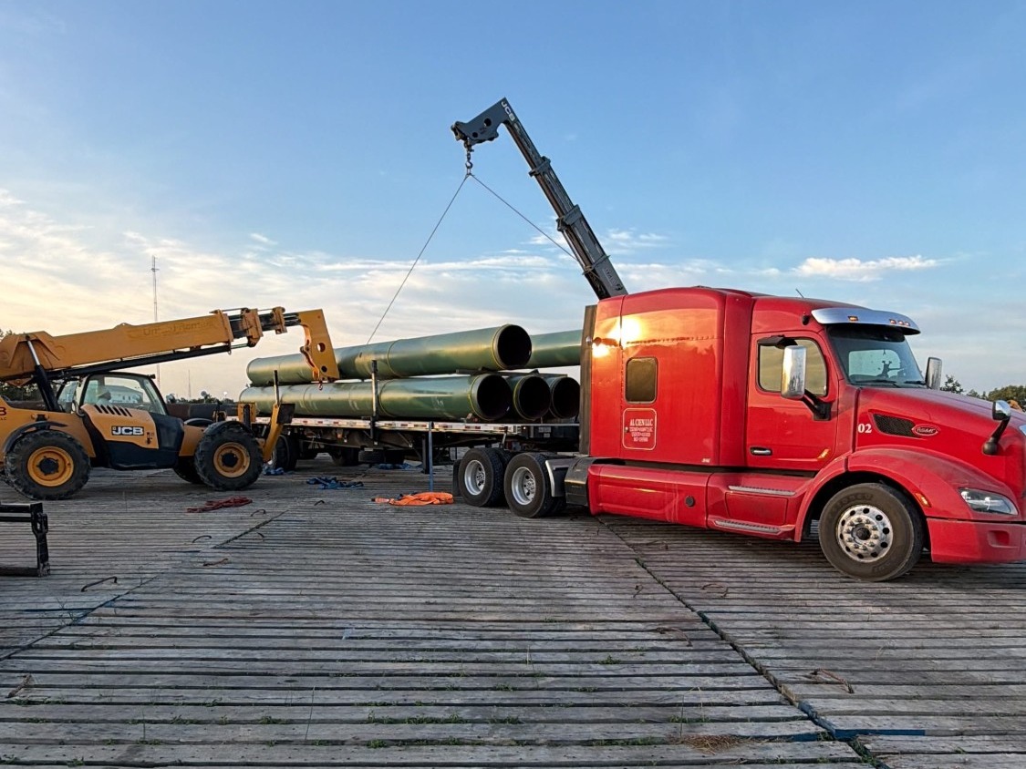 Knuckle boom crane and a JCB telehandler loading green coated large-diameter line pipe onto a flatbed trailer on a timber board road under a low sun