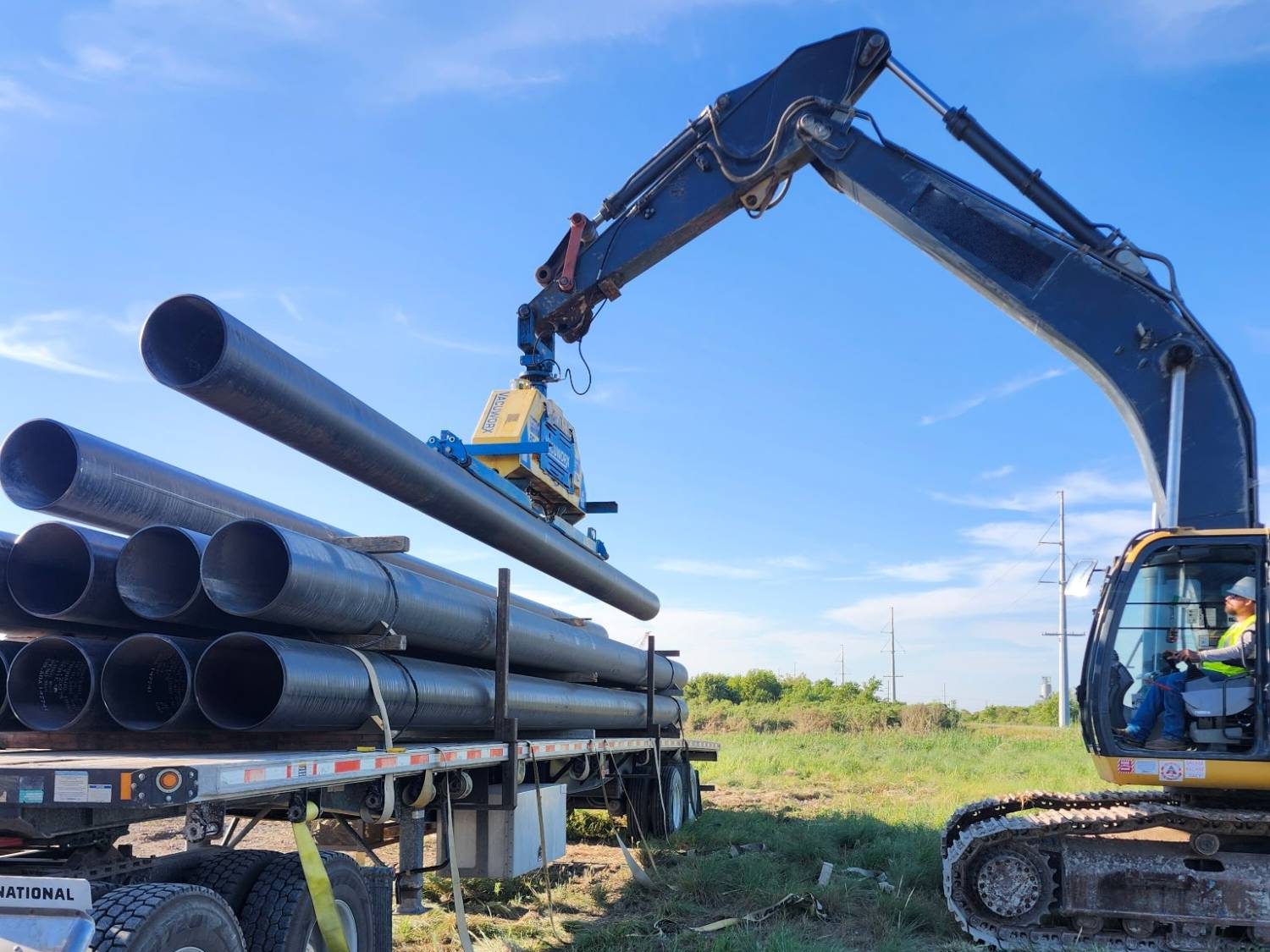 Excavator with a vacuum lifter setting a joint of large-diameter black steel pipe onto a loaded flatbed trailer in an open grass field