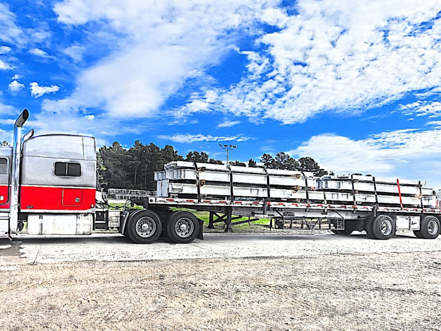 Red tractor with a silver sleeper pulling a flatbed loaded with strapped white fabricated structural steel, parked on a gravel lot under a bright sky