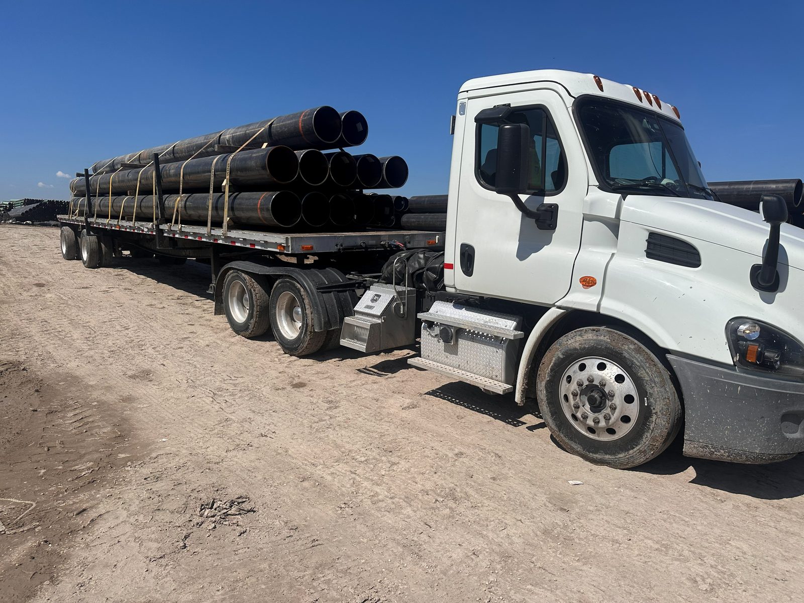 White day cab tractor pulling a flatbed loaded with strapped large-diameter black steel pipe at a pipe yard, more pipe stacked in the background