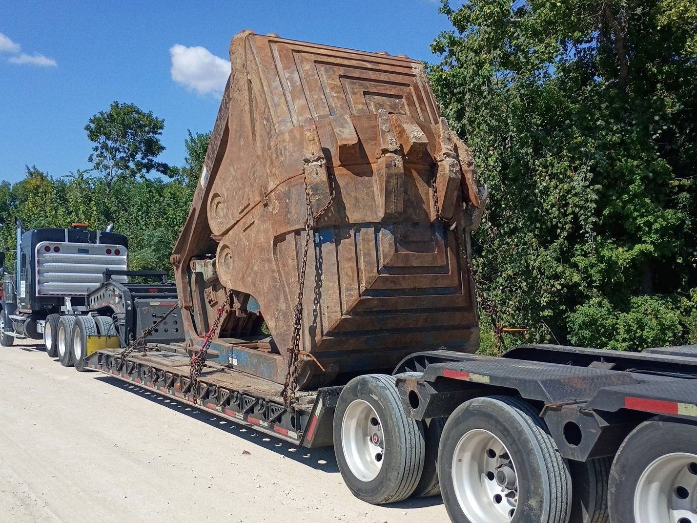 Lowboy trailer with a huge rusted excavator rock bucket chained to the deck, black tractor ahead, on a white gravel road lined with trees