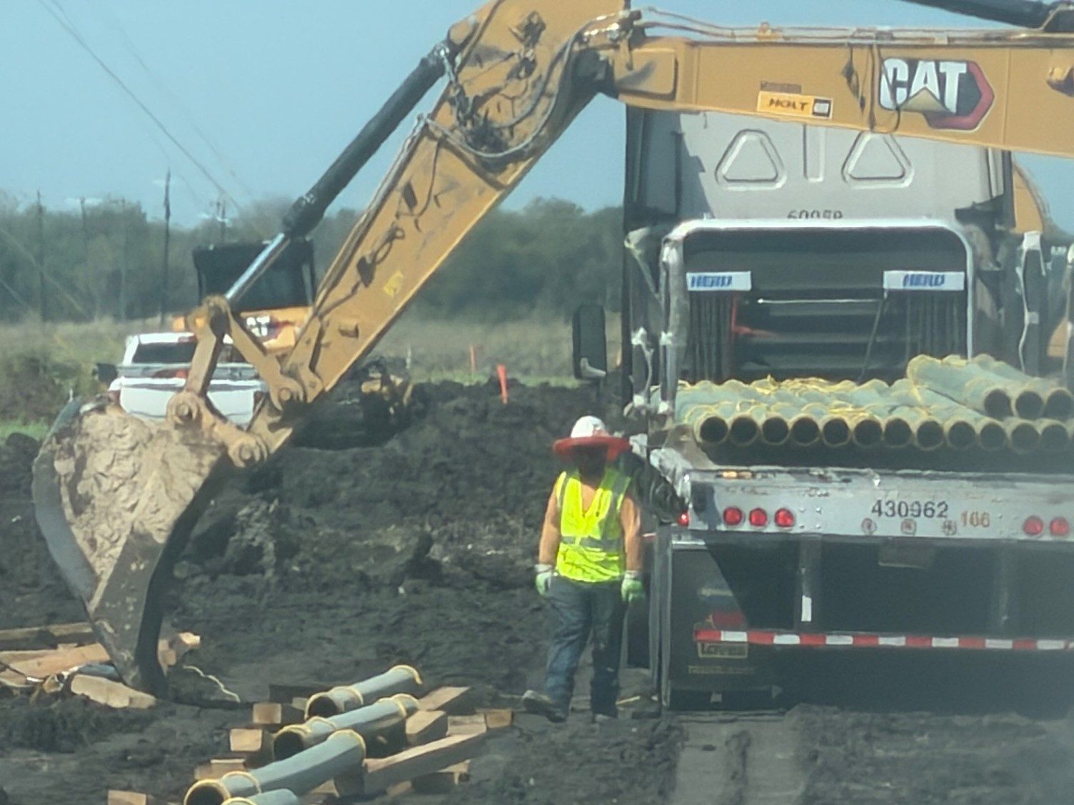 Excavator unloading a flatbed of green coated line pipe on a pipeline right-of-way, joints already strung out on timber skids, a worker in a hi-vis vest beside the trailer