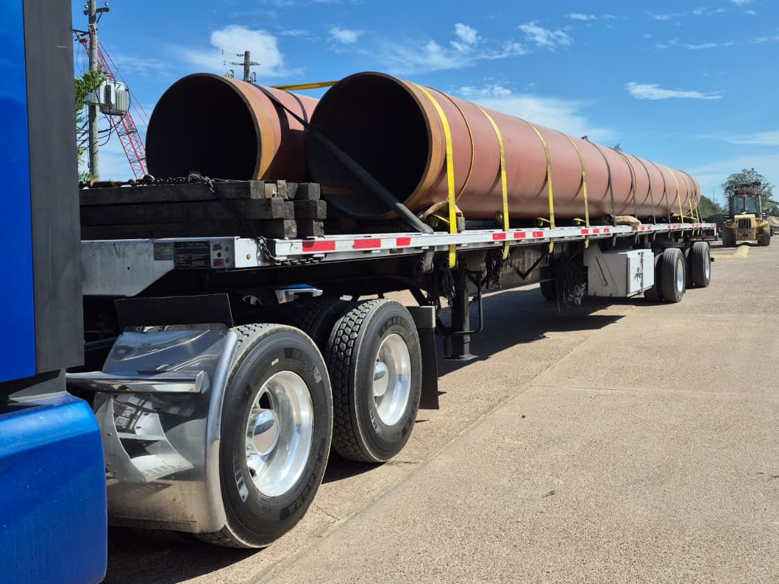 Blue tractor pulling a flatbed loaded with two lines of large-diameter red primed steel pipe, strapped down behind stacked timber dunnage, in a pipe yard