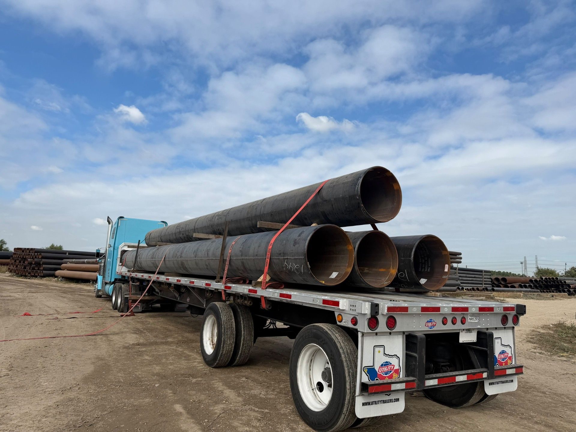 Light blue tractor pulling a flatbed loaded with four joints of large-diameter black steel pipe, three on the deck and one on timber dunnage above them, strapped down in a pipe yard, seen from the back of the trailer