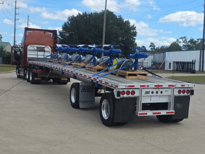 Red tractor pulling a flatbed loaded with a row of large valves fitted with blue electric actuators, each on its own pallet and strapped down, seen from behind the trailer in an industrial park