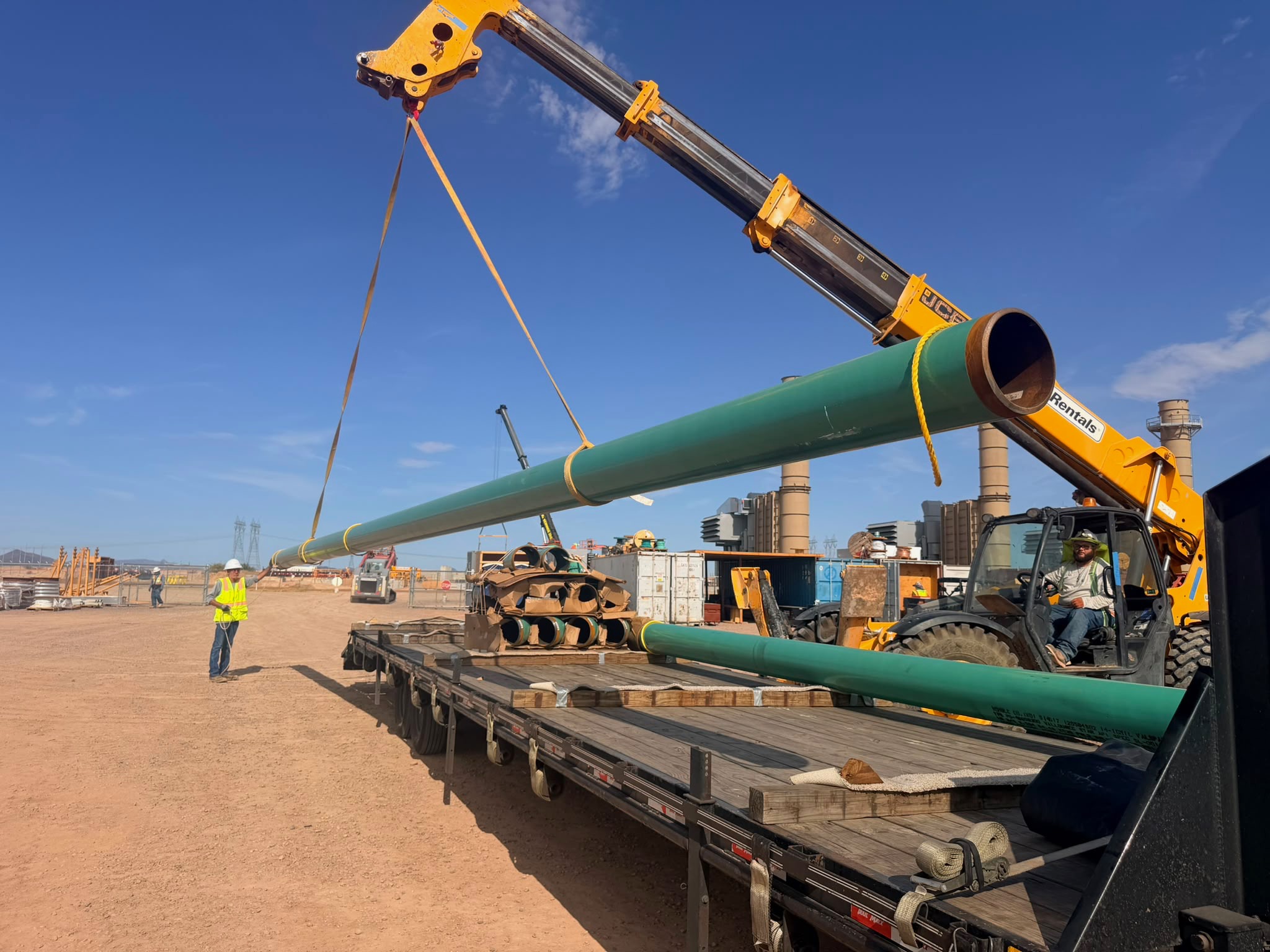 Crane loading large green pipe sections onto a flatbed trailer at a job site